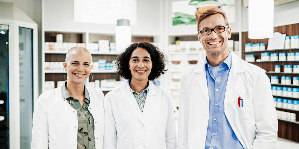Three dentists stand smiling in a room full with shelves full of medication